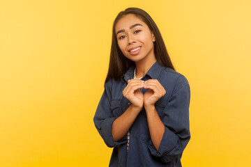 Portrait of amiable girl in denim shirt making heart shape gesture and smiling with kind friendly expression, showing love care, generosity symbol. indoor studio shot isolated on yellow background