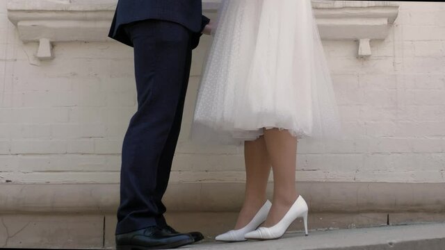 The legs of the bride and groom are standing opposite each other. In the background is a white brick building. Sunny summer day.