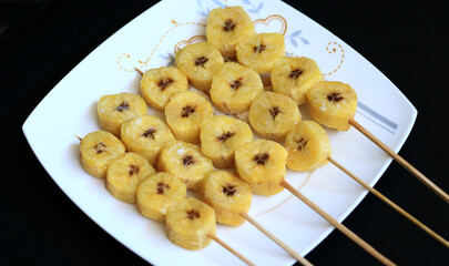 Banana satay on white plate, black background.