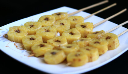 Banana satay on white plate, black background.