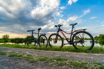 Naklejka premium Leisure activity in nature with two mountain or touring bicycles equipped with luggage compartment near a lake with reflections of clouds and reeds at sunset. Beautiful blue sky above a country road.