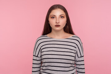 Portrait of serious calm young woman in striped sweatshirt looking at camera with unsmiling earnest expression, feeling confident focused self-assured. indoor studio shot isolated on pink background