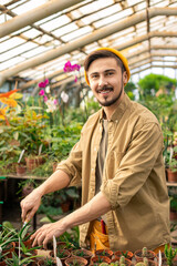 Portrait of happy young man in hat standing at counter with young plants in pots and planting garden in greenhouse