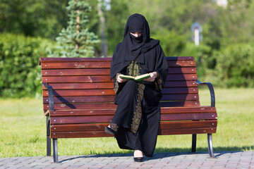 Arab woman in black national dress is reading a book in a city park A migrant from the Middle East...