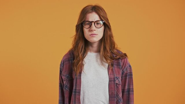 Ginger teenage girl in checkered shirt and glasses. She raising hands, asking why, looking upset and annoyed. Posing on orange background. Close up