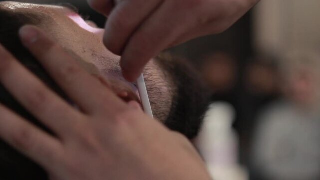 Barber shaves client’s beard with a straight razor blade in a barbershop, close-up.