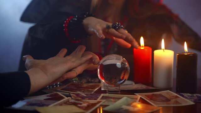 Young witch fortune teller reading fortune lines on male palm or hand, mystic esoteric or paranormal chiromancy concept. Future prediction with crystal ball and burning candles in dark room. Symbolism