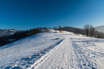 Fototapeta na wymiar Winter on Cienkow in Beskid Slaski mountains in Poland with snow covered road and meadows, trees, hills on the background and clear sky
