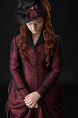 A young Victorian woman in a dark red and black ensemble against a black backdrop