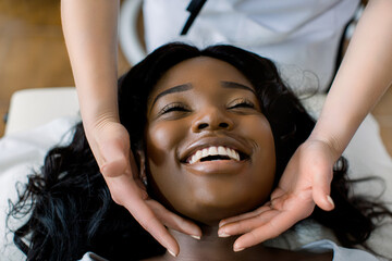 Close-up of hands of cosmetologist woman making special face massage to young pretty African...