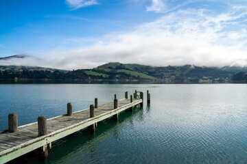 Fototapeta na wymiar Wooden pier and sailboat at Akaroa harbor, New Zealand. Clear sky with a few white clouds and mist. Symbol for relaxation, wealth, leisure activity. Panorama format.