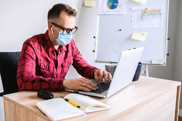 Young concentrated man works in the office while pandemic situation. An office worker sits at the office desk wearing medical mask with a laptop and typing on it, flipchart on the background