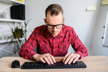 Webcam view of young concentrated man in casual plaid shirt at the office, he is typing on the keyboard and looking on it with a serious look on his face.