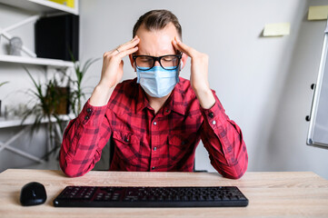 A guy wearing face mask in office sits at the desk with headache. Symptoms of the illness