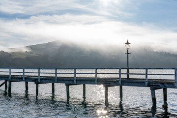 Fototapeta premium Wooden pier and sailboat at Akaroa harbor, New Zealand. Clear sky with a few white clouds and mist. Symbol for relaxation, wealth, leisure activity. Panorama format.