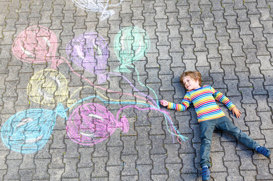 Little happy blond kid having fun with colorful air balloons picture drawing with colorful chalks. Creative leisure for children outdoors in summer, celebrating birthday. Chalk on asphalt