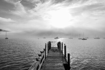 Fototapeta premium Wooden pier and sailboat at Akaroa harbor, New Zealand. Clear sky with a few white clouds and mist. Symbol for relaxation, wealth, leisure activity. Panorama format.