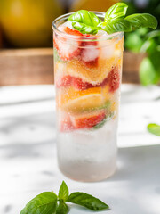 Fresh Summer Homemade lemonade with orange slices, strawbereies, basil and ice in glass on a white table.