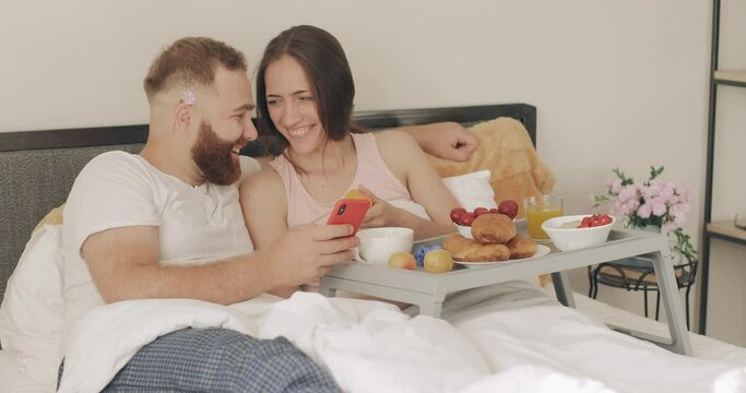 Happy young couple spending time and laughing in bed. Man and woman looking at smartphone screen and talking while having breakfast in early morning. Concept of leisure and relationship.