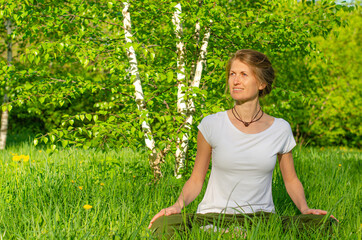 Young blond woman in white top practicing yoga in beautiful nature. Meditation in morning sunny day in the park with trees.