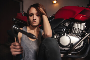 Fototapeta na wymiar A young girl sits in a garage on a background of a motorcycle. The girl holds a tool and looks at him with an incomprehensible look. The girl does not know how to use the tool