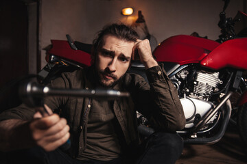 Fototapeta na wymiar A young man sits in a garage on a background of a motorcycle. A man holds an instrument and looks at him with an incomprehensible look. The man does not know how to use the tool