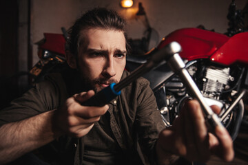 A young man in a garage holds a tool and does not know what to do with it. Close-up photo. Motorcycle on background