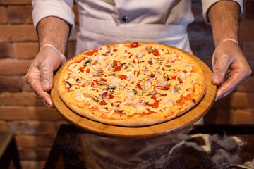 Chef in gloves holds appetizing pizza on the wooden stand, tablet. Pizza is made of shrimps, onion, paprica, cheese sauce. 