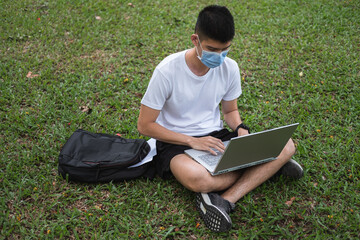 asian young man wearing mask is using laptop for social media in nature park.male student is working on notebook while sitting alone in garden keep social distancing in summer due to quarantine time.