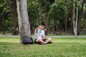 Obraz premium asian young man is looking at mask while sitting alone in park for social distancing after quarantine and stay safe. teenager hold mask in garden in summer.