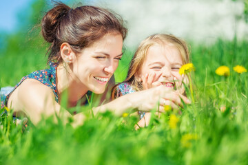 Young woman and her daughter resting on green summer grass with blooming yellow dandellions