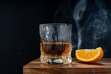 A glass of whiskey together with orange slice on wooden bar counter against dark background and smoke
