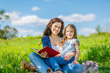 Mother and daughter enjoy reading book sitting on green meadow in summer park