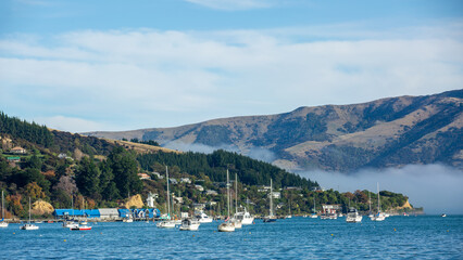 Fototapeta premium Blue sky, white clouds, mist & aquamarine seas at Akaroa Harbor. Anchored are fishing boats & sailboats, along the Port at Akaroa, Canterbury, New Zealand.