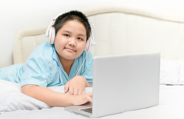 Obese schoolboy lying and wearing headphones to learn online from a laptop