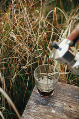 Alternative coffee brewing in travel. Pouring fresh hot coffee from geyser coffee maker into glass cup in sunny warm light in rural countryside herbs. Atmospheric moment. Vertical image