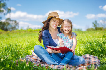 Naklejka na ściany i meble Mother and daughter enjoy reading book sitting on green meadow in summer park