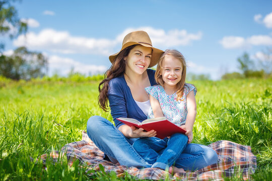 Mother and daughter enjoy reading book sitting on green meadow in summer park