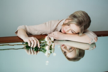 A beautiful girl with light hair is sitting next to a mirror in a room with a cold shade. Fashion shoot for a women's clothing brand