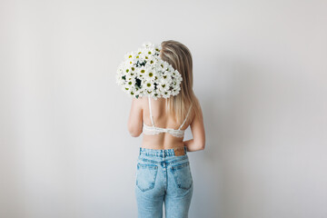 A beautiful girl with a covered face with light hair in her underwear holds a bouquet of daisies. A festive bouquet of daisies in the focus of the lens. A gift for women's day or mother's Day.