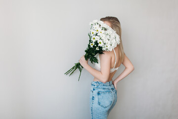 A beautiful girl with a covered face with light hair in her underwear holds a bouquet of daisies. A festive bouquet of daisies in the focus of the lens. A gift for women's day or mother's Day.