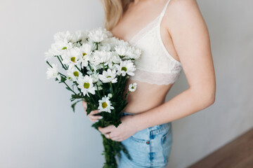 A beautiful girl with a covered face with light hair in her underwear holds a bouquet of daisies. A festive bouquet of daisies in the focus of the lens. A gift for women's day or mother's Day.