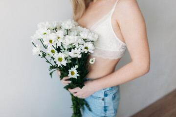 A beautiful girl with a covered face with light hair in her underwear holds a bouquet of daisies. A festive bouquet of daisies in the focus of the lens. A gift for women's day or mother's Day.