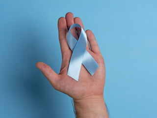 Close-up male hand holding blue prostate cancer awareness ribbon on a bright blue background....