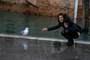 horizontal photo of a young woman serving a seagull standing on the edge of a venetian canal