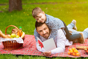 Happy Father Day. Cute child hugging his dad, while he is reading greeting card on picnic in forest