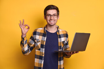 Portrait of positive smart confident cool man in eyeglasses holding laptop, making okay hand...