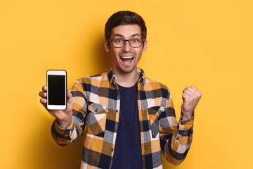 Excited millennial man isolated on yellow studio background, showing smartphone with blank copy space, yes hand gesture, text message, client use cellular, read great news, win lottery on mobile app