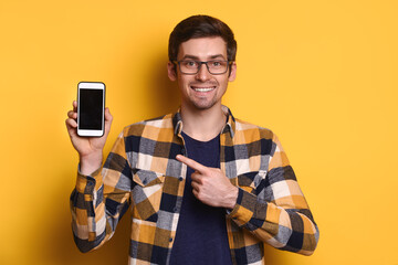 Cheerful caucasian man in glasses pointing with finger at blank smartphone display in his hand, smiling, looking at the camera, chatting, advice, advertising. Isolated on yellow background. Copy space