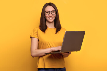 Young cute woman in eyeglasses is smiling happily, holding laptop isolated over yellow background, looking at the camera. Copy space. Promotion concept